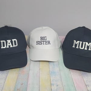 Puede incluir: Tres gorras de béisbol. Dos son de color azul marino con texto bordado blanco "DAD" y "MUM". La tercera gorra es blanca con "BIG SISTER" bordado en gris. Todas tienen viseras curvas.