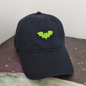 Puede incluir: Gorra de béisbol azul marino con visera curva. La gorra presenta un logotipo de murciélago bordado en verde neón en la parte delantera. La gorra está hecha de tela suave y tiene un pequeño botón en la parte superior.