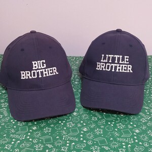 May include: Two navy blue baseball caps. One cap has the text "BIG BROTHER" embroidered in white, and the other has "LITTLE BROTHER" in white. The caps are on a green patterned surface.
