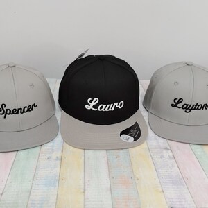 May include: Three personalized baseball caps, one black with white lettering that says "Lauro", one gray with white lettering that says "Layton", and one gray with white lettering that says "Spencer".