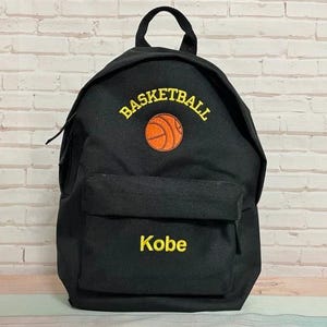 Puede incluir: Mochila negra con un bolsillo frontal y un asa superior. La palabra "BASKETBALL" está bordada en amarillo sobre un gráfico de baloncesto naranja. El nombre "Kobe" está bordado en amarillo debajo del bolsillo frontal.