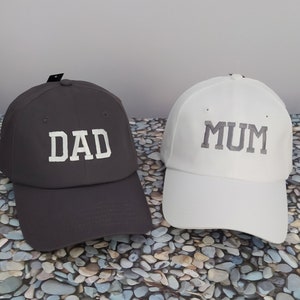 A pair of two baseball hats with unstructured front panel, one black hat embroidered with the text DAD with white thread and one white hat embroidered with the text MUM with white thread. Nice gift for couples, who are going to become parents