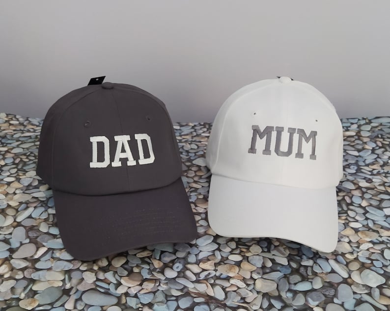 A pair of two baseball hats with unstructured front panel, one black hat embroidered with the text DAD with white thread and one white hat embroidered with the text MUM with white thread. Nice gift for couples, who are going to become parents