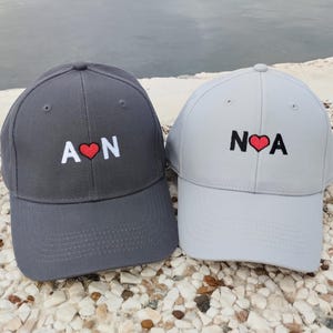 Peut inclure: Deux casquettes de baseball, une grise foncée et une grise claire, sont présentées. Chaque casquette porte des initiales brodées avec un symbole de cœur rouge. La casquette grise foncée affiche "A ❤️ N" et la grise claire "N ❤️ A".