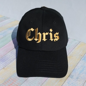 Pode incluir: Boné de beisebol preto com o nome "Chris" bordado em letras douradas. O boné tem uma aba curva e uma coroa estruturada. O fundo é uma superfície clara com um padrão multicolorido.