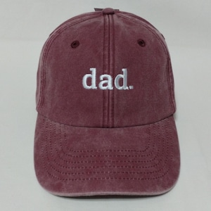 Könnte beinhalten: Eine weinrote Baseballkappe mit dem Wort "dad." in Weiß gestickt. Die Kappe hat einen gebogenen Schirm und einen verstellbaren Riemen. Der Hut ist aus weichem, abgenutztem Material.