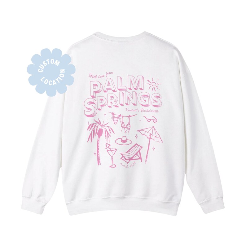 May include: White sweatshirt with a pink graphic design on the back. The design includes the words "Palm Springs" with beach-themed illustrations like a palm tree, bikini, and beach umbrella. The text "Kendall's Bachelorette" is also included.