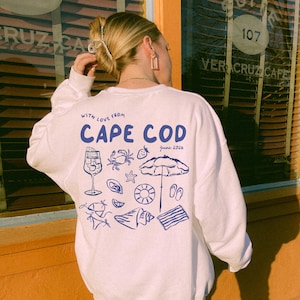 May include: A white long-sleeve sweatshirt featuring a blue graphic on the back with the text "WITH LOVE FROM CAPE COD" and "june 2026". The design includes illustrations of a drink, a crab, a strawberry, a starfish, an umbrella, and a shell.