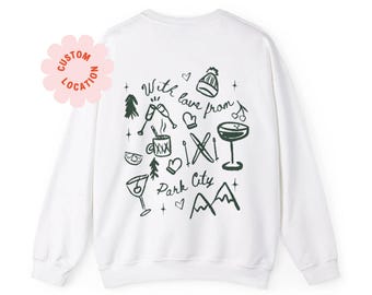 May include: White sweatshirt with a green graphic design that says "With love from Park City" and includes illustrations of winter activities like skiing, a mug of hot chocolate, and a cocktail.