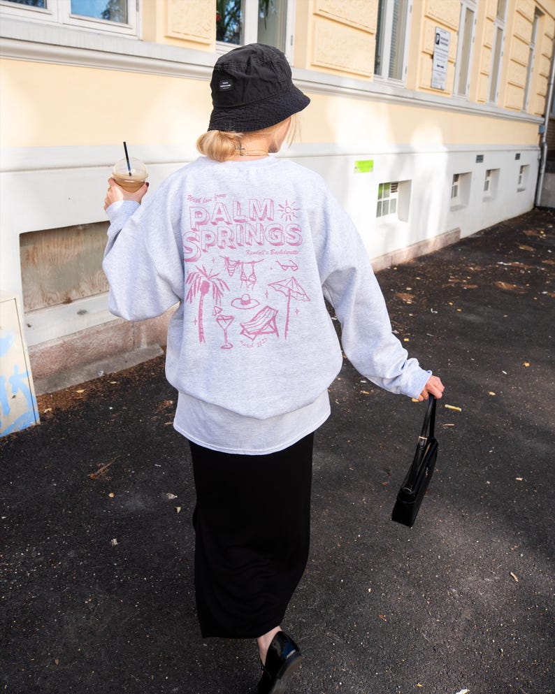 May include: A light gray sweatshirt with a pink graphic that says "Palm Springs" with illustrations of a palm tree, cocktail, and beach chair. The person is wearing a black bucket hat and a long black skirt.