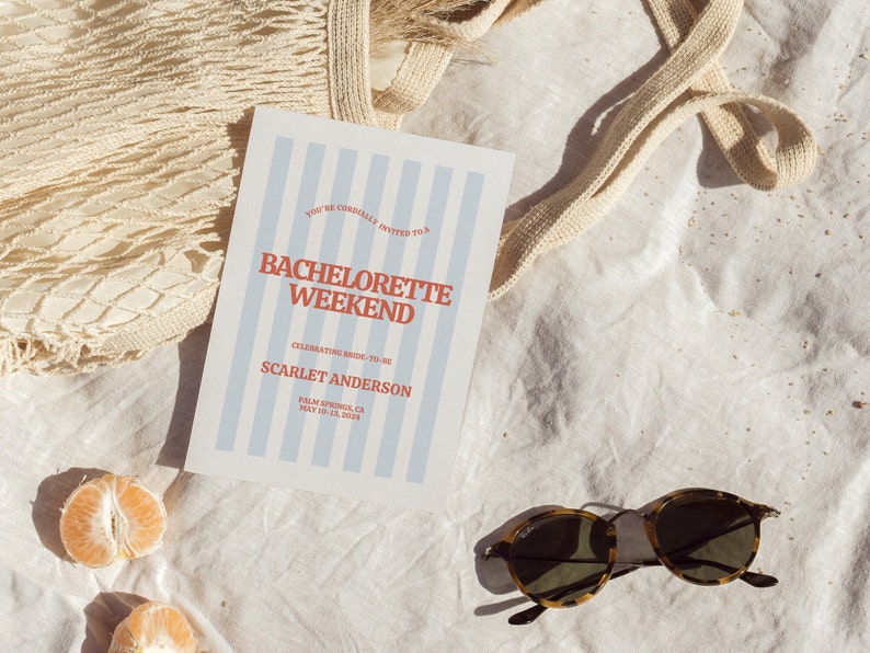 May include: A white card with blue stripes and red text announcing a bachelorette weekend for Scarlet Anderson in Palm Springs, CA from May 10-13, 2024. The card is lying on a white fabric surface with a tan mesh bag and two halved mandarin oranges.