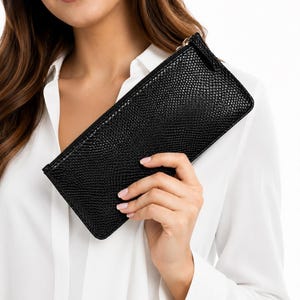 May include: A slim, rectangular black clutch wallet featuring a textured, lizard-skin embossed pattern. The accessory has a top zip closure with a small pull tab and a structured, elongated shape, designed for holding essentials in a compact form.