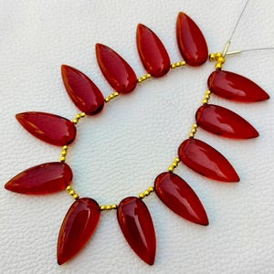 May include: A strand of red gemstone beads, each bead is shaped like a teardrop. The beads are strung on a gold wire with small gold beads between each gemstone.