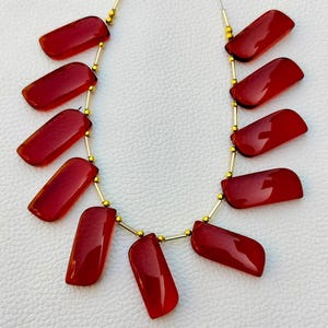 May include: A strand of red gemstone beads strung on a gold chain. The beads are rectangular and have a smooth, polished finish.