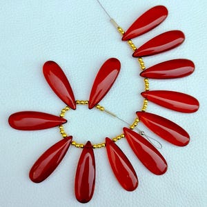 May include: A strand of red gemstone beads strung on a gold wire. The beads are teardrop shaped and have a glossy finish.