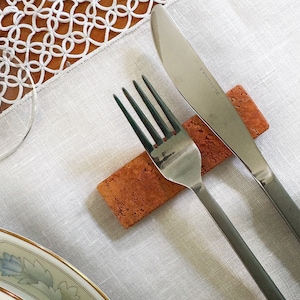 Puede incluir: Un reposacubiertos rectangular de corcho texturizado en color marrón rojizo, colocado horizontalmente. Un tenedor de acero inoxidable plateado y un cuchillo a juego con la inscripción STAINLESS STEEL en la hoja descansan sobre la pieza de corcho.