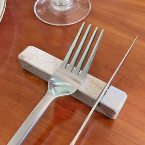 May include: A silver fork and knife resting on a light grey marble utensil holder. The holder has a light brown veining pattern.