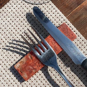 May include: A silver fork and knife are placed on a woven placemat with a red and white rectangular piece of food between them.