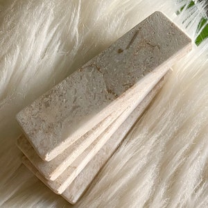 Questo poggiaposate è in marmo, con una tonalità beige chiara e una finitura leggermente ruvida. La superficie presenta leggere intrusioni e imperfezioni che conferiscono un aspetto rustico. La forma è rettangolare con bordi totalmente smussati.