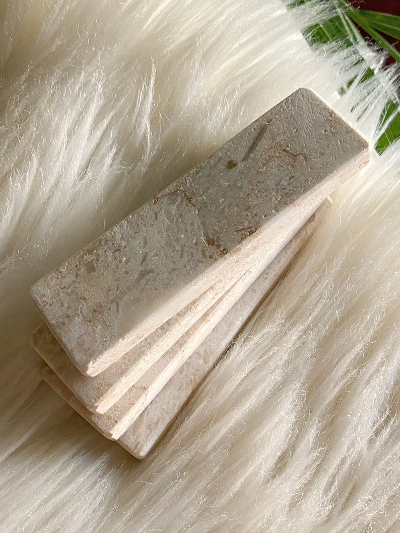 Questo poggiaposate è in marmo, con una tonalità beige chiara e una finitura leggermente ruvida. La superficie presenta leggere intrusioni e imperfezioni che conferiscono un aspetto rustico. La forma è rettangolare con bordi totalmente smussati.