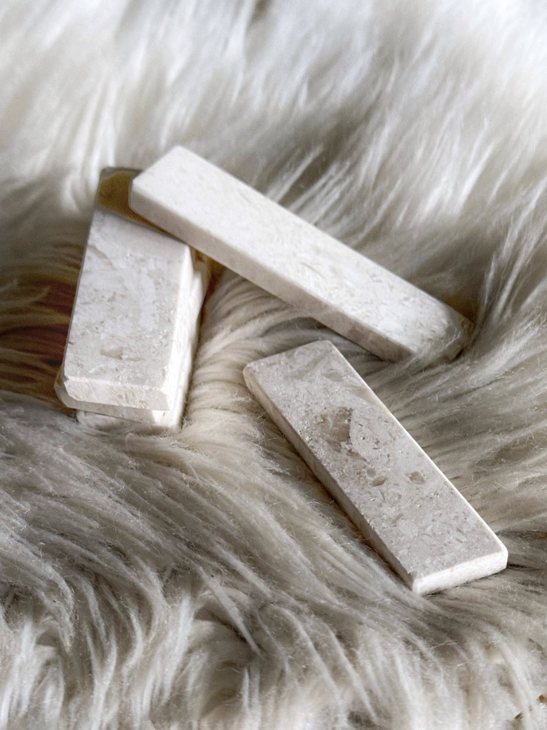 Questo poggiaposate è in marmo, con una tonalità beige chiara e una finitura leggermente ruvida. La superficie presenta leggere intrusioni e imperfezioni che conferiscono un aspetto rustico. La forma è rettangolare con bordi totalmente smussati.