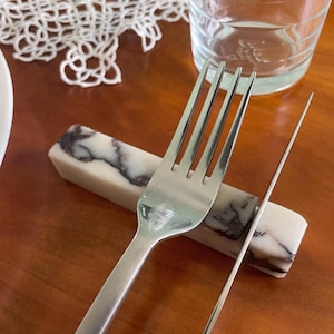 May include: A rectangular chopstick rest made of white stone with dark grey veining. The piece features a smooth, polished finish and a solid, block-like shape. It is positioned horizontally to support a stainless steel fork and a thin metal knife.