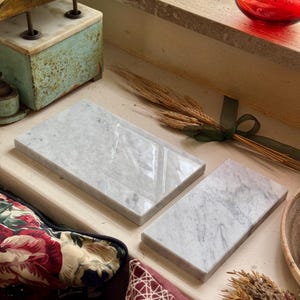 May include: Two rectangular white marble slabs with grey veining. The larger slab is positioned to the left, while the smaller slab is placed to the right. Both pieces feature a smooth, polished surface and straight, clean edges.