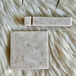 Puede incluir: Un posavasos cuadrado de piedra beige con una textura natural moteada y un soporte rectangular de piedra beige que presenta una ranura vertical estrecha para sostener objetos. Ambas piezas exhiben una apariencia de piedra uniforme y porosa.