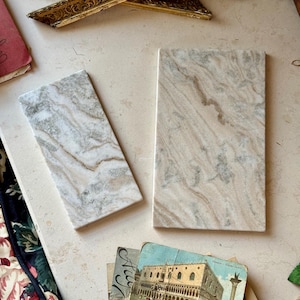 Può includere: Due lastre rettangolari in marmo con finitura lucida, caratterizzate da motivi vorticosi di venature bianche, beige e grigie. Le lastre sono posizionate fianco a fianco, mostrando una texture in pietra naturale con superfici lisce e piatte.