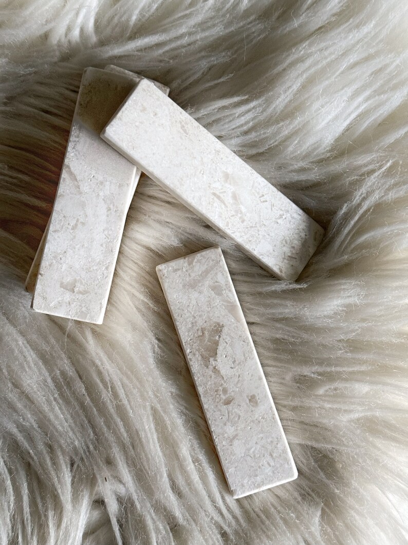 Questo poggiaposate è in marmo, con una tonalità beige chiara e una finitura leggermente ruvida. La superficie presenta leggere intrusioni e imperfezioni che conferiscono un aspetto rustico. La forma è rettangolare con bordi totalmente smussati.