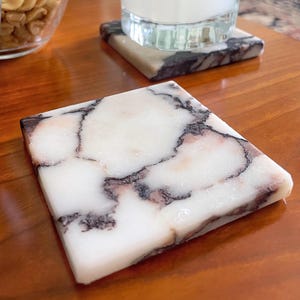 Può includere: Un sottobicchiere quadrato in pietra bianca con venature marrone scuro e nere che attraversano l'intera superficie. Il sottobicchiere ha una finitura liscia e lucida e presenta motivi organici irregolari simili alla pietra di marmo.