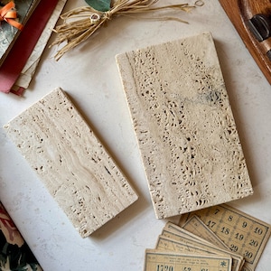 Op de afbeelding: Twee rechthoekige beige travertijn onderzetters met een ruw, getextureerd oppervlak. De onderzetters liggen op een wit oppervlak met een stapel vintage speelkaarten met nummers en een ster symbool.