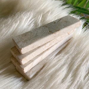 Questi poggiaposate sono in marmo, con una tonalità beige chiara e una finitura leggermente ruvida. La superficie presenta leggere intrusioni e imperfezioni che conferiscono un aspetto rustico. La forma è rettangolare con bordi totalmente smussati.
