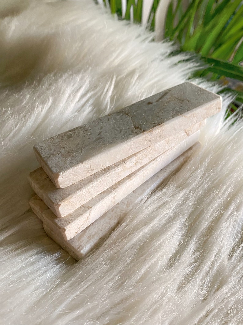 Questi poggiaposate sono in marmo, con una tonalità beige chiara e una finitura leggermente ruvida. La superficie presenta leggere intrusioni e imperfezioni che conferiscono un aspetto rustico. La forma è rettangolare con bordi totalmente smussati.