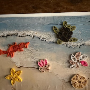 Op de afbeelding: Een papier-quilling kunstwerk met een strandscène, bestaande uit een bruine zeeschildpad, een oranje krab, een gele zeester en twee bloemachtige schelpvormen in roze, witte en beige tinten op een bedrukte achtergrond van een kustlijn.