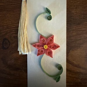 May include: A rectangular light-colored wooden bookmark featuring a paper quilling design of a six-petaled pink flower with a yellow center. The flower is accented by green paper quilled vines and leaves. A cream-colored tassel is attached at the top.