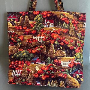 Op de afbeelding: Een bruine tote bag met een herfstthema print met rode schuren, pompoenen en bomen. De print is op een donkerbruine achtergrond.