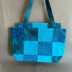 以下が含まれることがあります： 正方形のデザインの青いパッチワークトートバッグ。バッグには2本の長いストラップが付いています。