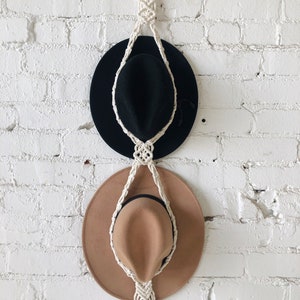 May include: A white macrame hat rack with two hats hanging on it. The top hat is black and the bottom hat is brown.