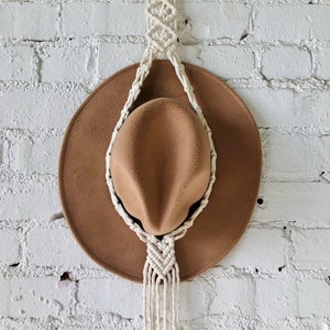 May include: A tan fedora hat hangs from a cream-colored macrame hat hanger. The hat has a wide brim and a rounded crown. The macrame hanger is attached to a wooden ring at the top and is suspended against a white brick wall.
