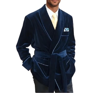 May include: A navy blue velvet robe with light blue trim and a matching belt. The robe has a shawl collar, pockets, and a pocket square. The model is wearing a white shirt and yellow tie. This robe is a luxurious addition to any wardrobe.