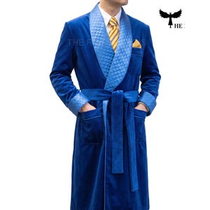 May include: A royal blue velvet robe featuring a quilted light blue shawl collar and matching quilted cuffs. The robe includes a tie belt, two front pockets, and a chest pocket containing a yellow pocket square. A yellow and blue striped tie is visible.