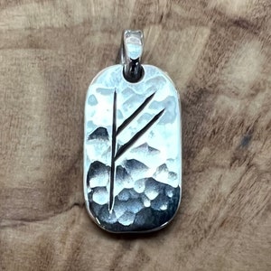 Może przedstawiać: Srebrny wisiorek z młotkowanym wykończeniem i wygrawerowanym symbolem runy. Runa to prosty wzór z dwoma pionowymi liniami.