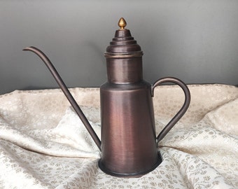 May include: A copper oil and vinegar dispenser with a spout and a decorative top. The dispenser is sitting on a white fabric with a floral pattern.
