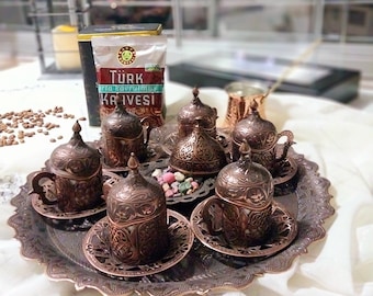 Pode incluir: Um conjunto de seis xícaras de café turco de cobre adornadas e pires em uma bandeja decorativa. As xícaras têm desenhos florais intrincados e estão dispostas em círculo. A bandeja é de metal e tem uma borda festonada. As xícaras estão cheias de café e há pequenos doces na bandeja.