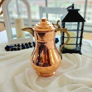 May include: A hammered copper pitcher with a brass handle and spout. The pitcher has a traditional design and is perfect for serving beverages.