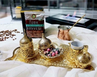 May include: A gold-colored Turkish coffee set with a tray, two cups, and a pot. The set is on a white tablecloth with a bag of Turkish coffee in the background. The coffee bag has the text "Türk Orta Kavrulmuş Kahvesi" on it.