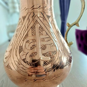 May include: A copper-colored pitcher with an ornate, engraved design. The pitcher has a decorative handle and a wide base. The etched pattern covers the body of the pitcher, creating a textured surface. The pitcher is likely used for serving beverages.
