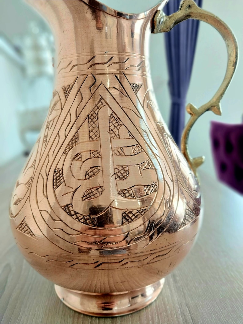 May include: A copper-colored pitcher with an ornate, engraved design. The pitcher has a decorative handle and a wide base. The etched pattern covers the body of the pitcher, creating a textured surface. The pitcher is likely used for serving beverages.