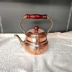 May include: A copper tea kettle with a hammered finish and a wooden handle. The kettle has a spout and a lid. It is sitting on a white tablecloth.